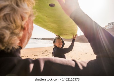 couple of seniors at the beach holding a surftable on their heads to prepare to go surfing with wetsuits - vacation and freetime to have fun with a surf lesson - Powered by Shutterstock