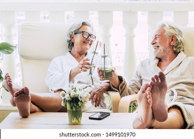 Couple Of Seniors Or Mature People In A Resort Spa In Their Hotel Or House Clinking With Their Cocktail Or Drink - Having Fun With Feet On The Table Looking Between They