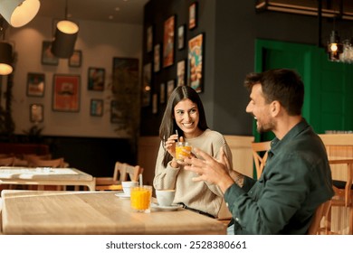 Couple shares a joyful moment at a stylish café, sipping coffee and enjoying bright orange juice while laughing together, surrounded by a welcoming atmosphere - Powered by Shutterstock