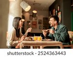 Couple shares laughter and delicious food while sitting at a wooden table in a bright cafe, surrounded by greenery and other patrons enjoying their meals