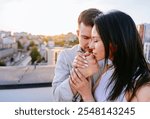 Couple sharing intimate moment on rooftop as sun sets behind city skyline. Man and woman embrace with hands clasped, affection and connection. Calmness of evening setting enhances romantic atmosphere.