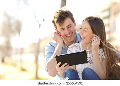 Couple sharing music and singing with a tablet sitting in a bench in an urban park - Powered by Shutterstock