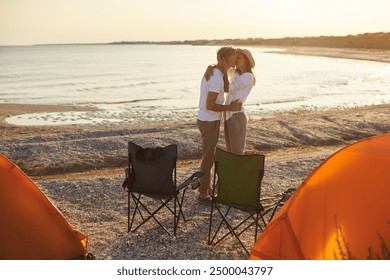 A couple sharing a romantic kiss at their beachside campsite with orange tents during sunset, creating a perfect and intimate moment by the sea. - Powered by Shutterstock