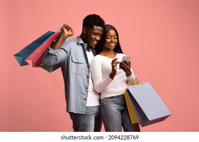 Couple of shopaholics. Happy black millennial man and woman holding shopping bags, enjoying their new goods, shopping online together, using smartphone, pink studio background, copy space
