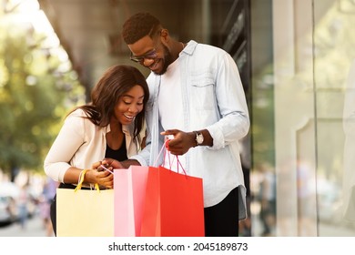 Couple Of Shopaholics. Smiling African American family of two holding shopping bags, looking inside, excited about their new purchases, standing outside near mall centre. Gifts, Retail And Joy