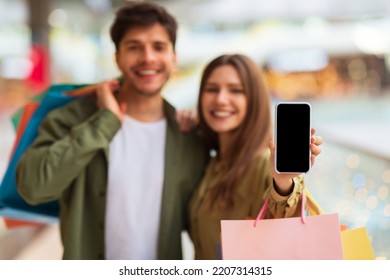 Couple Showing Cellphone Blank Screen Advertising Shopping Application Holding Shopper Bags Posing Standing In Modern Hypermarket Indoors. Shallow Depth, Mockup