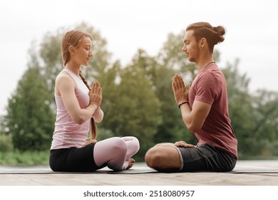 A couple sits cross-legged in a serene outdoor environment, practicing yoga while focusing on mindfulness. They are positioned facing each other, hands in a prayer gesture. - Powered by Shutterstock