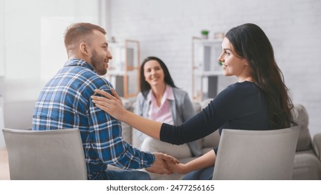 A couple sits facing each other in a therapy session, holding hands. The woman is smiling at her partner, while the man looks happy and relaxed. A lady counselor sits behind them - Powered by Shutterstock