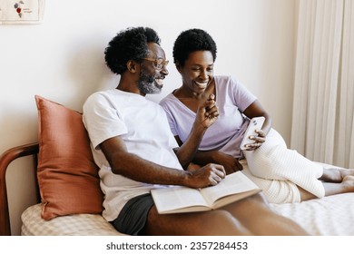 Couple sits on a bed, enjoying time together at home. Husband holds a book while the wife smiles, using her phone with him. Mature couple using technology in a relaxed and romantic moment. - Powered by Shutterstock