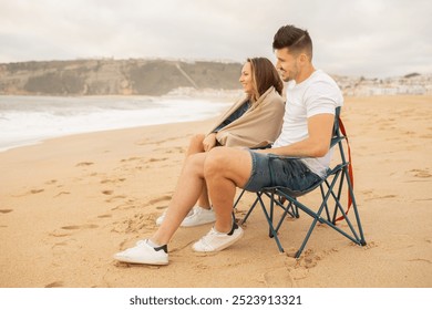 A couple sits on chairs on a sandy beach, wrapped in a blanket as they enjoy the ocean view. The calm waves and distant cliffs create a tranquil atmosphere for relaxation. - Powered by Shutterstock