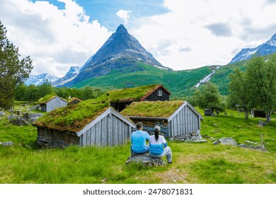 A couple sits on a rock, enjoying the scenic view of traditional Norwegian cabins with grass roofs, set against a backdrop of majestic mountains in Norway. Innerdalen valley in Trollheimen mountains - Powered by Shutterstock