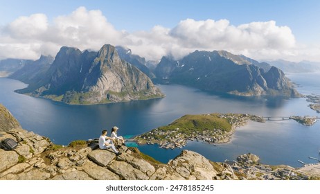 A couple sits on a rocky cliff overlooking a stunning Norwegian fjord. Lush green mountains and a small village dot the landscape. a diverse couple hiking the Reinebringen mountain, Lofoten, Norway - Powered by Shutterstock