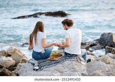 A couple sits on a rocky shore sharing a meal beside calming waves. - Powered by Shutterstock