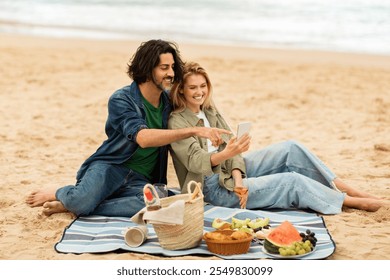 A couple sits on the sand, enjoying a picnic spread with fruits and snacks. They laugh and share moments while looking at a smartphone, surrounded by a calm beach at sunset. - Powered by Shutterstock