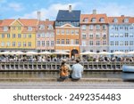 A couple sits on a stone wall overlooking a bustling Copenhagen canal, taking in the vibrant facade of historic buildings and the lively atmosphere of the waterfront. Copenhagen Denmark 