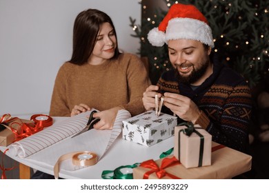 couple sits table wrapping presents Christmas tree background. woman cutting wrapping paper scissors. man ties ribbon wrapped box. concept: christmas gift wrapping  - Powered by Shutterstock