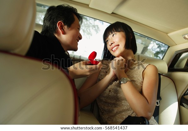 Couple sitting in backseat of car, man\
holding ring box, woman smiling with hands\
clasped