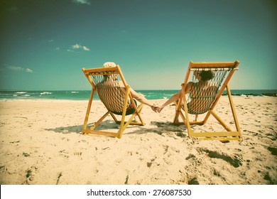 Couple sitting in beach chairs and holding hands on a tropical beach  - retro style background