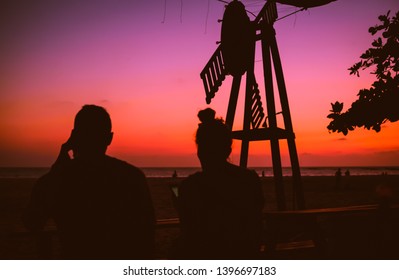 A Couple Sitting At A Beach Shack Distracted By Their Phone And Missing Out On A Beautiful Orange And Pink Sunset In Front Of Them.