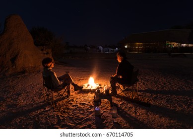 Couple Sitting At Burning Camp Fire In The Night. Camping In The Desert With Wild Elephants In Background. Summer Adventures And Exploration In The African National Parks.
