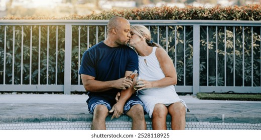 Couple sitting by a pool, enjoying a moment together. They are sharing a kiss, holding drinks, and relaxing by the poolside, enjoying the sunny day. Diverse couple kissing while sitting by pool. - Powered by Shutterstock