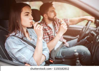 Couple Is Sitting In Car. Guy Is Driving And Eating Candy Bar. Girl Is Looking Straight And Biting A Piece Of Bar As Well. They Are Safety Locked With Seatbelt