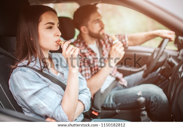 Couple is sitting in car. Guy is\
driving and eating candy bar. Girl is looking straight and biting a\
piece of bar as well. They are safety locked with\
seatbelt