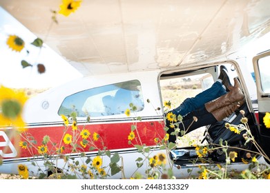 A couple sitting in the cockpit of a small airplane with their feet up on the dashboard. The airplane is in a field of sunflowers and the photo is taken from the side of the plane with the door open.