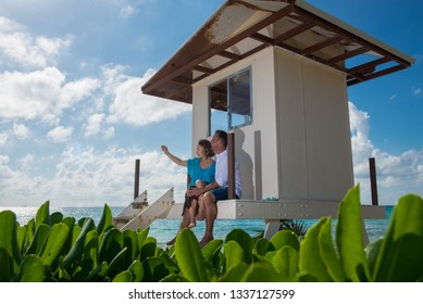 Couple Sitting Cousy At The Porch Of Tiny House House Or Beach Watcher's House In Cancun. Women Points At Something At The Sky. Turquoise Water Behind Them, And Green Leafs In Front.Backlight Picture.