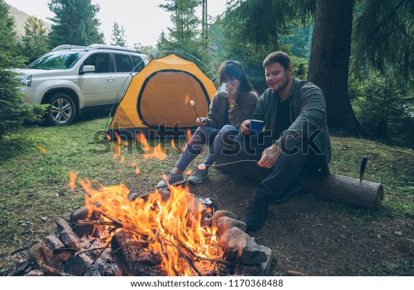 couple sitting\
near camp fire and drinking tea and telling stories . tent and suv\
on background. union with\
nature
