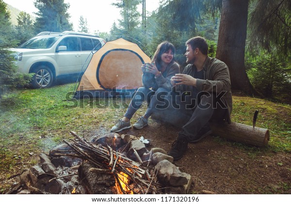couple sitting\
near camp fire and drinking tea and telling stories . tent and suv\
on background. union with\
nature