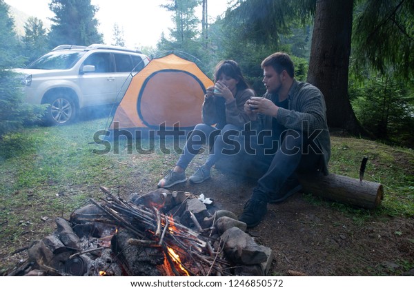 couple sitting near camp\
fire and drinking tea. tent and suv on background. union with\
nature