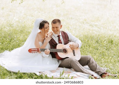 A couple is sitting on a blanket in a grassy field, with a guitar in front of them. The man is wearing a red vest and the woman is wearing a white wedding dress - Powered by Shutterstock