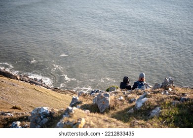 Couple Sitting On A Cliff-side Ledge Overlooking The Sea. Sightseers By The Beach.