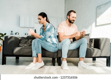 Couple Sitting On Couch, Facing Opposite Direction And Using Smartphones At Home