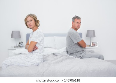 Couple Sitting On Different Sides Of Bed Not Talking After Argument Looking At Camera In Bedroom At Home