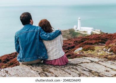 Couple Is Sitting On The Edge Of Cliff, South Stack Lighthouse On Background, Wales, Anglesey. Staycation And Travelling Around UK.