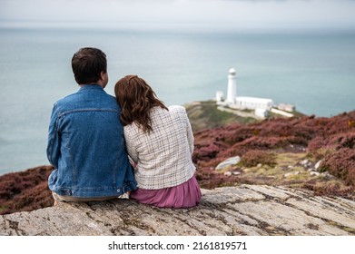 Couple Is Sitting On The Edge Of Cliff, South Stack Lighthouse On Background, Wales, Anglesey. Staycation And Travelling Around UK.