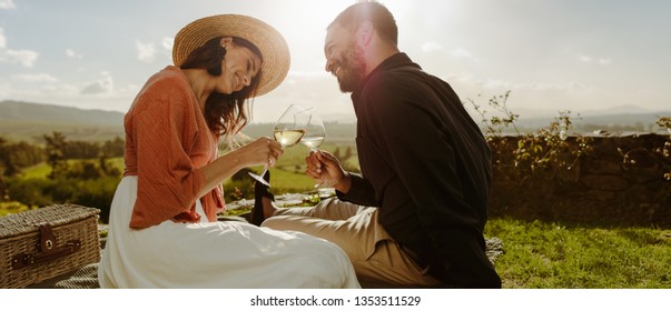 Couple sitting on grass in a vineyard toasting wine. Smiling woman in hat sitting with her boyfriend drinking wine and talking to him. - Powered by Shutterstock