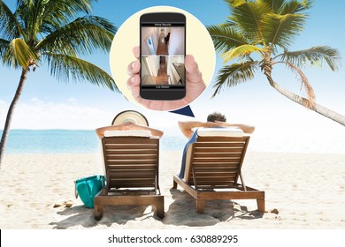 Couple Sitting On Lounge Chair At Beach With Home Security Cameras Viewed On Mobile Phone