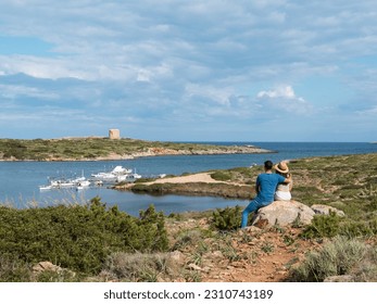 A couple sitting on a rock, observing a landscape of a natural bay with a small fishing port in the sea, with a historic tower in the background, on a sunny day with clouds in Menorca, Balearic Island - Shutterstock ID 2310743189