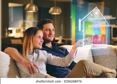 A Couple Sitting On The Sofa Controls All The Functions Of The House Such As Wi-fi, Heating, Lighting, Television Through Holography. Concept Of, Home Automation, Automations, Future, Technology.