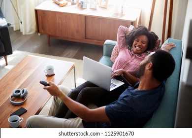 Couple Sitting On Sofa At Home Using Laptop Computer And Watching TV