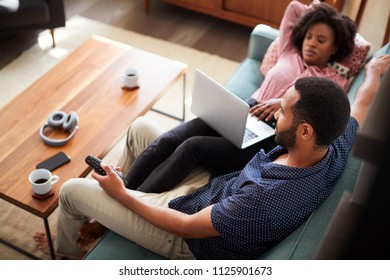 Couple Sitting On Sofa At Home Using Laptop Computer And Watching TV