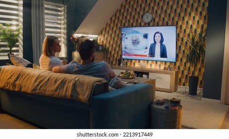 Couple sitting on sofa in living room, talking, watching TV news about poliovirus and water pollution. Man and woman spending leisure time on weekend at home together. TV live broadcasting. - Shutterstock ID 2213917449
