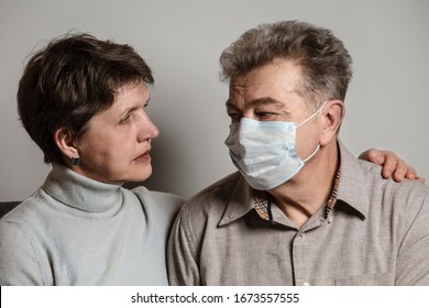 A Couple Sitting On A Sofa Wearing A Protective Mask. An Infectious Agent Protection Gear  Including A Mask. An Sick Older Man And Woman Wearing Protective Masks To Protect Against Virus. Coronavirus.