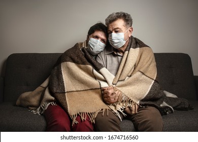 A Couple Sitting On A Sofa Wearing A Protective Mask. An Infectious Agent Protection Gear Including A Mask. An Sick Older Man And Woman Wearing Protective Masks To Protect Against Virus. Coronavirus.