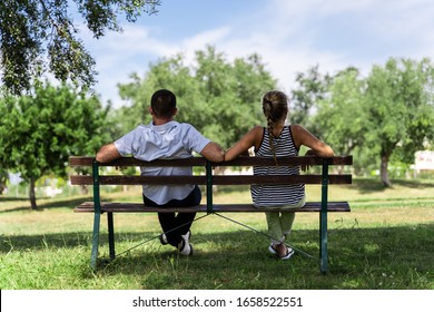 Couple sitting on a wooden bench under a tree looking forward to the horizon in the middle of a park with trees - Powered by Shutterstock