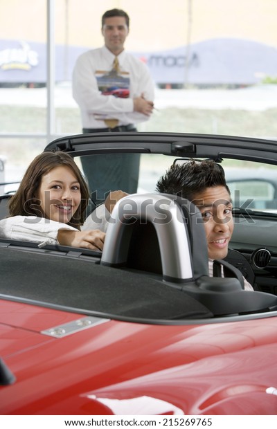 Couple sitting in red convertible in\
car showroom, salesman in background, smiling,\
portrait