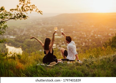 Couple Sitting At Summer Picnic With Hands Up And Watching Sunset At A Mountain, While Making A Toast With Wine During Golden Hour.